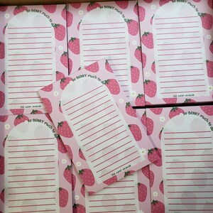 May include: Pink notepads with a strawberry pattern and the text "so BERRY much to do." Each notepad has a curved top and lined paper. The brand name "the violet veranda" is printed at the bottom of each notepad.