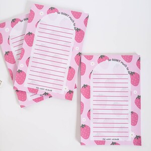 May include: Pink notepads with a strawberry pattern and the text "so BERRY much to do." Each notepad has a lined section for writing and the text "the violet veranda." Daisies are also printed on the notepads.