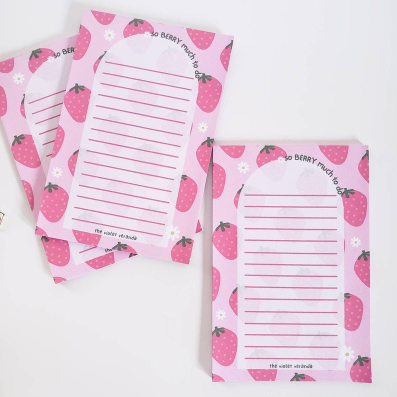 May include: Pink notepads with a strawberry pattern and the text "so BERRY much to do." Each notepad has a lined section for writing and the text "the violet veranda." Daisies are also printed on the notepads.