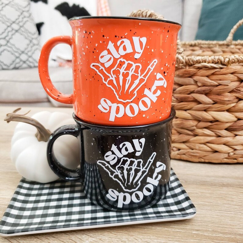 May include: Two ceramic mugs with black and white skeleton hand designs and the text "Stay Spooky". The mugs are stacked on top of each other. The top mug is orange with black speckles and the bottom mug is black with white speckles.