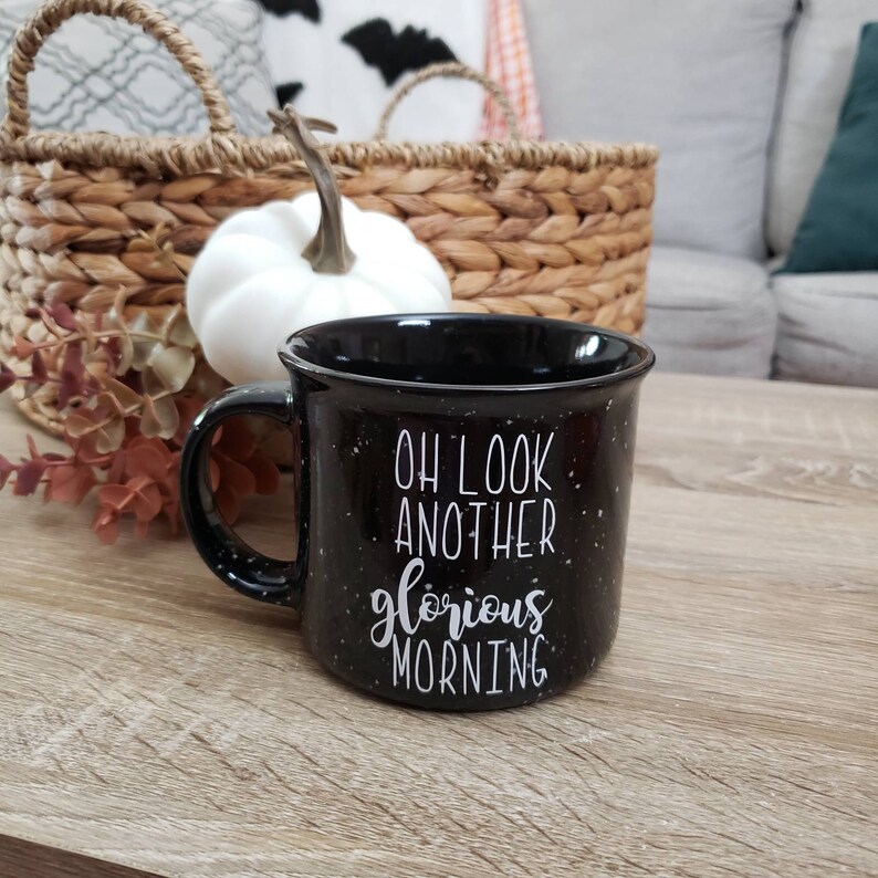 May include: Black speckled ceramic mug with the text "OH LOOK ANOTHER glorious MORNING" in white script. The mug has a black handle and a wide rim. The mug is sitting on a wooden surface.