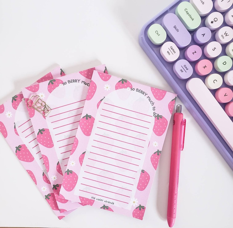 May include: Pink notepad with strawberry pattern and the text "So Berry Much to Do". A pink pen and a pastel keyboard are also in the image. The notepad has lined pages and a gold paperclip.