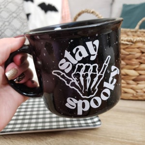 May include: Black ceramic mug with white speckled glaze. The mug has a white graphic of a skeleton hand making a shaka sign with the text "stay spooky" around it.