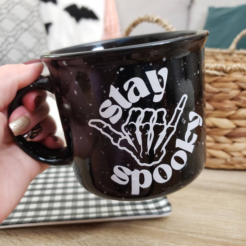 May include: Black ceramic mug with white speckled glaze. The mug has a white graphic of a skeleton hand making a shaka sign with the text "stay spooky" around it.