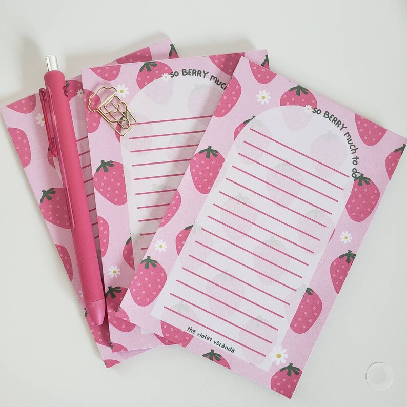 May include: Pink notepad with a strawberry pattern and the text "so BERRY much to do." A pink pen and gold paperclip are next to the notepad. The notepad has lined pages and the text "the violet veranda."