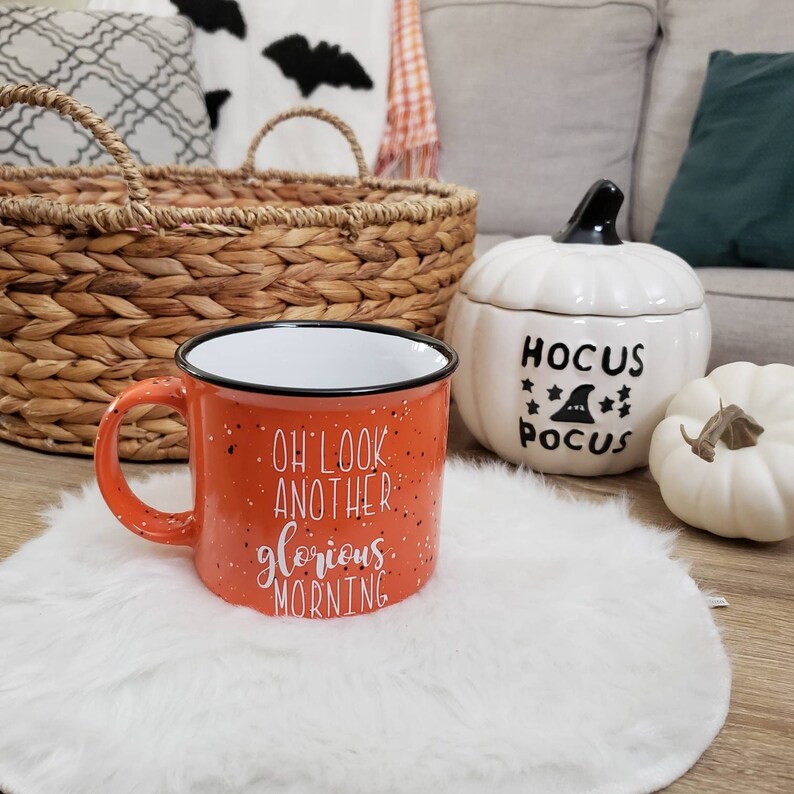 May include: An orange enamel mug with black speckles and the words "Oh look another glorious morning" in white. A white pumpkin-shaped container with "Hocus Pocus" text and a small white pumpkin are also visible.