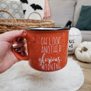 May include: An orange ceramic mug with black speckles and a black rim. The mug has the text "OH LOOK ANOTHER glorious MORNING" in white script. The mug is held by a person's hand.
