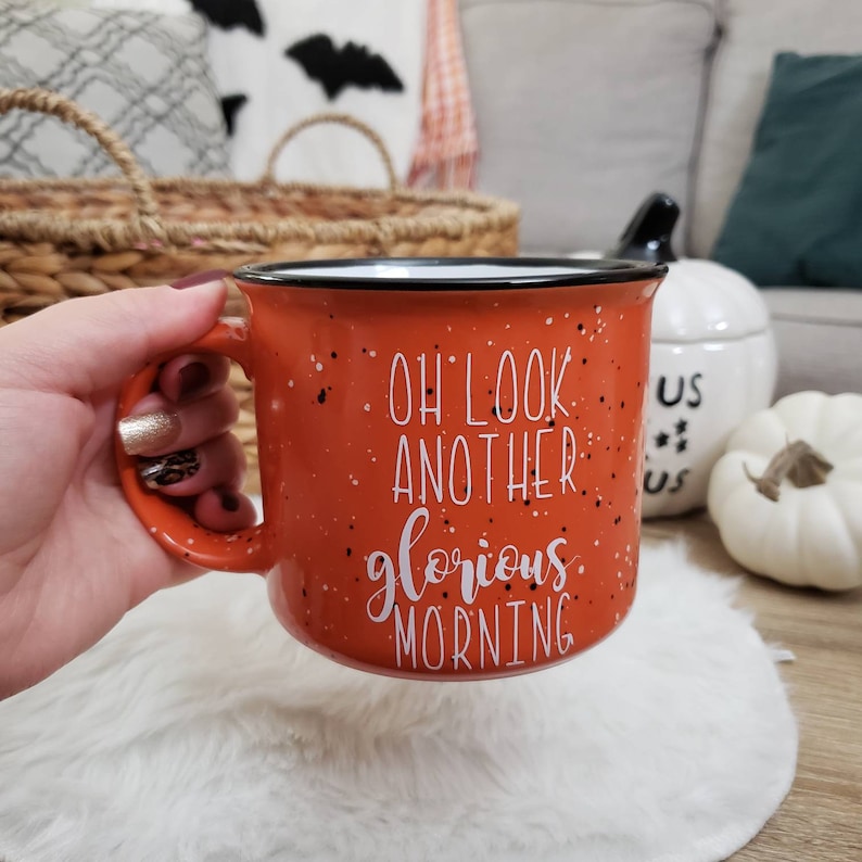 May include: An orange ceramic mug with black speckles and a black rim. The mug has the text "OH LOOK ANOTHER glorious MORNING" in white script. The mug is held by a person's hand.