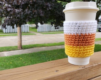 May include: A white coffee cup with a crocheted candy corn-colored cozy. The cozy is white, orange, and yellow.