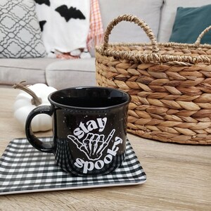 May include: Black coffee mug with white speckled glaze and the text "Stay Spooky" with a skeleton hand making a shaka sign. The mug is on a black and white checkered coaster.