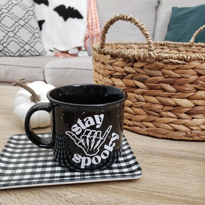 May include: Black coffee mug with white speckled glaze and the text "Stay Spooky" with a skeleton hand making a shaka sign. The mug is on a black and white checkered coaster.