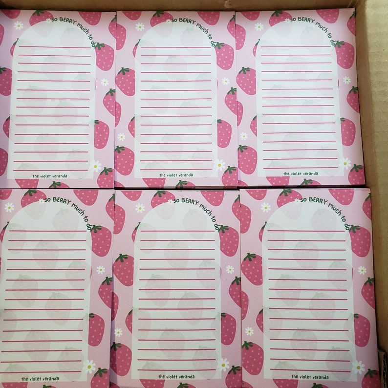 May include: A stack of pink notepads with a strawberry pattern and the text "so BERRY much to do." Each notepad has a white arched section with lined paper and the text "the violet veranda."