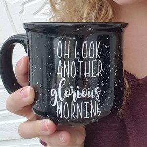 May include: Black speckled ceramic mug with the text "OH LOOK ANOTHER glorious MORNING" in white script. The mug has a black handle and is held by a person. The mug is a camp style mug.