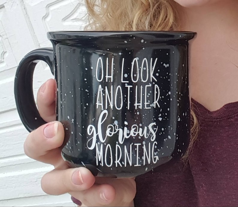 May include: Black speckled ceramic mug with the text "OH LOOK ANOTHER glorious MORNING" in white script. The mug has a black handle and is held by a person. The mug is a camp style mug.