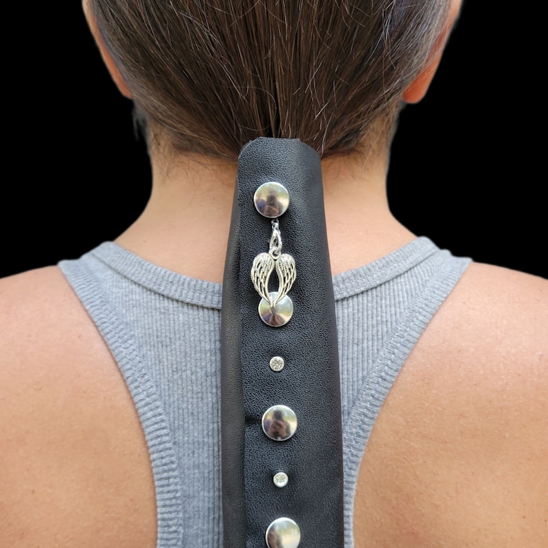 May include: Black leather hair accessory with silver-colored studs and a dangling charm featuring angel wings. The accessory is attached to the back of a person's head, with a gray tank top visible.