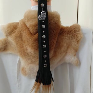 May include: Black leather hair accessory with silver studs and a decorative heart charm. The accessory features a fringe at the bottom and is designed to hold a ponytail. The hair accessory is shown on a mannequin.