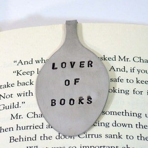 May include: A silver spoon bookmark with the words "LOVER OF BOOKS" stamped on it. The bookmark is laying on a page of a book.