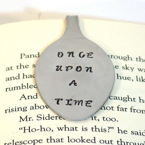 May include: A silver spoon bookmark with the words "Once Upon A Time" engraved on it. The bookmark is lying on a page of a book.