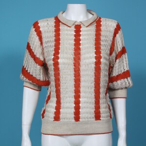Pode incluir: Uma camisa de malha creme de mangas curtas com listras verticais laranja. A camisa tem gola e punhos e bainha canelados. As mangas são ligeiramente bufantes. A roupa é exibida em um manequim branco sobre um fundo azul.
