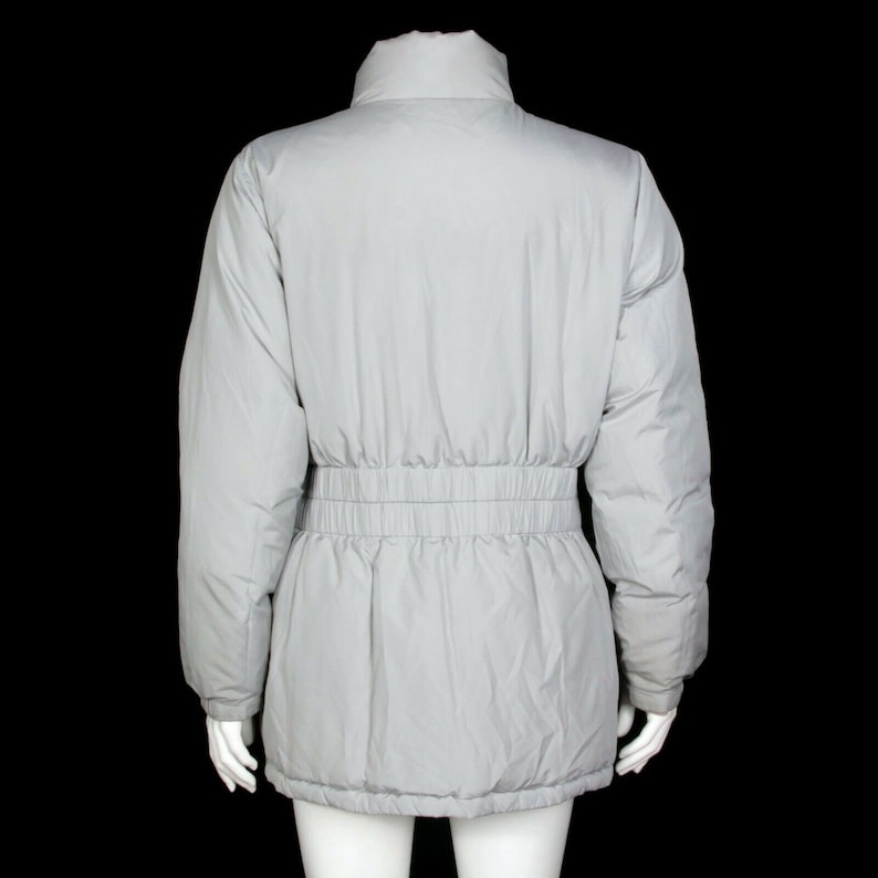 May include: A light gray puffer jacket with a cinched waist. The jacket has a high collar and long sleeves.
