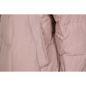 May include: A close-up of a pale pink puffer jacket with a quilted design. The jacket has a zipper closure and a pocket on the left side.