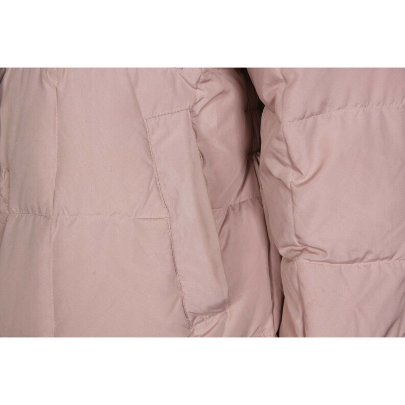 May include: A close-up of a pale pink puffer jacket with a quilted design. The jacket has a zipper closure and a pocket on the left side.