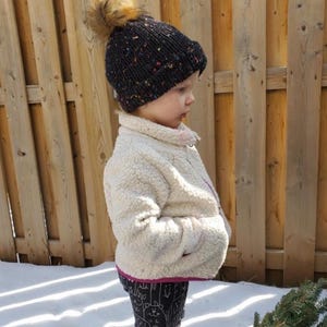 May include: A child wearing a black and white speckled knit beanie with a brown faux fur pom pom, a cream colored sherpa jacket with a pink zipper, and black and white patterned leggings.