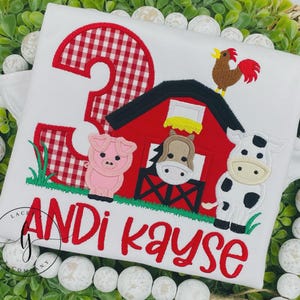 May include: White t-shirt with a red gingham number 3 and a barnyard scene. The design includes a red barn, a pig, a horse, a cow, and a rooster. The name "Andi Kayse" is embroidered in red.
