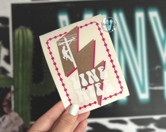 May include: A white sticker with a pink border and a lightning bolt design. The lightning bolt is brown and pink, with the words "LINE LIFE" in white. A silhouette of a lineman is on the left side of the lightning bolt.