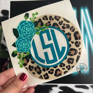 May include: A round sticker featuring a teal monogram within a white circle, surrounded by a leopard print border. Teal floral accents and the text "Vinylosity" are also present. The sticker is held by a hand.