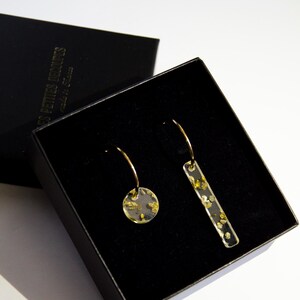 Peut inclure: Une paire de boucles d'oreilles dorées dans une boîte noire. L'une est un petit cercle, l'autre un rectangle. Les deux sont transparentes avec des paillettes dorées. La boîte est ouverte, révélant les boucles d'oreilles.