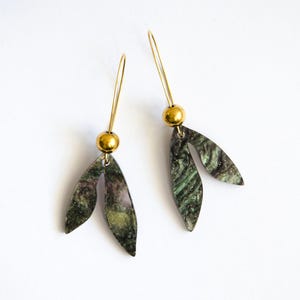 May include: A pair of dangle earrings with a leaf-like design. Each earring features a gold-colored post, a gold-colored ball, and a leaf-shaped charm with green, purple, and gold tones. The earrings are set against a white background.