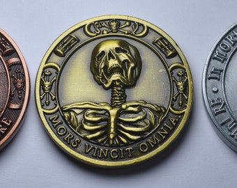 May include: Three round metal coins with skull imagery. The first coin is copper colored and reads "Memento Vivere". The second coin is gold colored and reads " Mors Vincit Omnia". The third coin is silver colored and reads "In Morte Ultima Veritas".