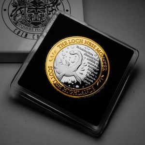 May include: A gold and silver coin with a depiction of the Loch Ness Monster and the text "The Loch Ness Monster Scottish Highlands".