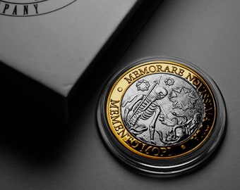 Op de afbeelding: Een zilveren munt met een gouden rand toont een skeletfiguur dat een speer en een schedel vasthoudt. De munt heeft de inscriptie "MEMENTO MORI" en "MEMORARE NOBIS" rond de rand.