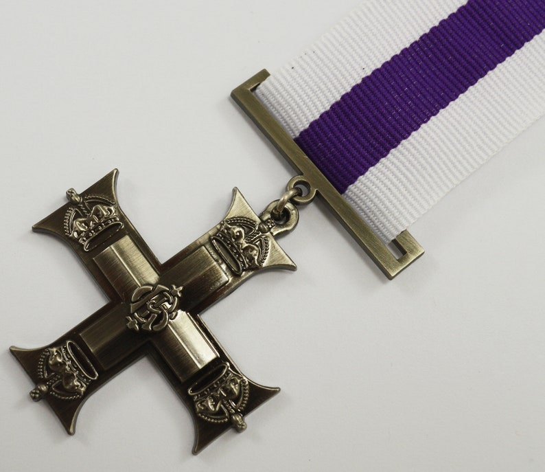 Pu&ograve; includere: Una croce di bronzo con quattro corone e un emblema centrale, appesa a un nastro bianco e viola.