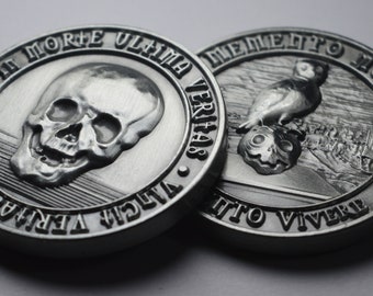 Op de afbeelding: Twee zilveren munten met Latijnse inscripties. De eerste munt toont een schedel met de tekst "IN MORTE ULTIMA VERITAS". De tweede munt toont een vogel die op een schedel zit met de tekst "MEMENTO MORI VITO VIVERE".
