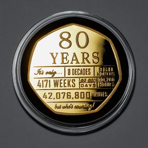 Puede incluir: Una moneda de color dorado con el número 80 y la palabra "YEARS" inscrita en ella. La moneda también incluye el texto "It's only... 8 DECADES | 960 MONTHS | 4171 WEEKS | 39,320 DAYS | 701,280 HOURS | 42,076,800 MINUTES but who's counting!"