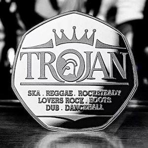 May include: A silver-colored, octagonal coin with the word "TROJAN" in large, bold letters. The coin also features a crown and a stylized warrior's helmet. The text "SKA. REGGAE. ROCKSTEADY. LOVERS ROCK. ROOTS. DUB. DANCEHALL." is printed around the edge of the coin.