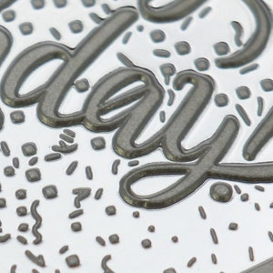 May include: A silver metal charm with the word "everyday" embossed in a dark gray color. The charm is decorated with a confetti-like pattern of small gray squares and lines.