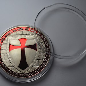 Op de afbeelding: Een zilveren en rode Tempeliersmunt met een rood kruis op een schild. De munt bevindt zich in een doorzichtig plastic doosje. De munt heeft de Latijnse zin "Non nobis domine, non nobis, sed nomini tuo da gloriam" gegraveerd rond de rand.