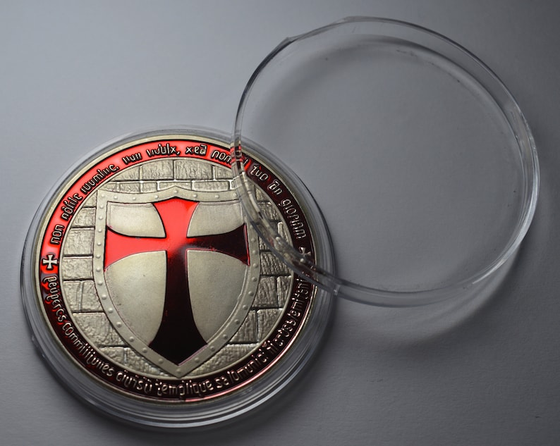 Op de afbeelding: Een zilveren en rode Tempeliersmunt met een rood kruis op een schild. De munt bevindt zich in een doorzichtig plastic doosje. De munt heeft de Latijnse zin "Non nobis domine, non nobis, sed nomini tuo da gloriam" gegraveerd rond de rand.