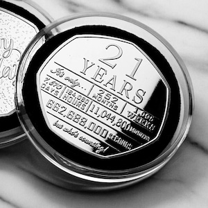 May include: A silver coin with a black background and the text "21 YEARS" in a white font. The coin also includes the text "It's only..." and a breakdown of the number of days, hours, months, weeks, minutes, and seconds in 21 years.