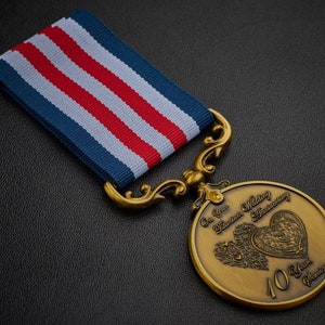 Può includere: Una medaglia dorata con un nastro blu, bianco e rosso. La medaglia presenta un design a forma di cuore con il testo "On Your Titanium Wedding Anniversary" e "40 Years Service".