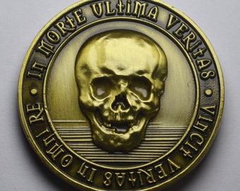 Op de afbeelding: Een gouden metalen munt met een schedel en gekruiste beenderen ontwerp. De munt heeft de Latijnse zin "In Morte Ultima Veritas" gegraveerd rond de rand.