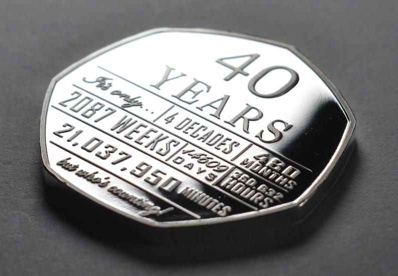 以下が含まれることがあります： 「40 YEARS」の文字と、週、日、分などの様々な時間の単位が刻まれた銀貨。 光沢のある反射面を持ち、暗い背景に配置されています。