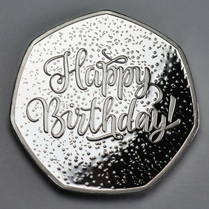 May include: A silver coin with a domed surface and a raised inscription that reads "Happy Birthday!" The coin has a speckled background.