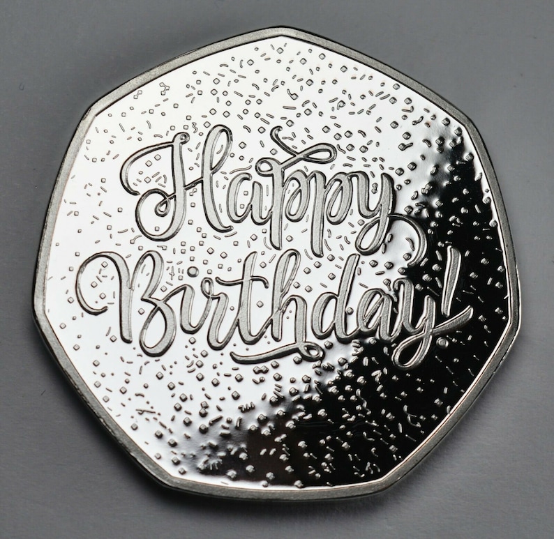 May include: A silver coin with a domed surface and a raised inscription that reads "Happy Birthday!" The coin has a speckled background.
