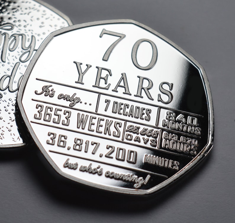 May include: A silver coin with the inscription "70 YEARS" and a breakdown of the number of weeks, days, hours, and minutes in 70 years. The inscription reads "It's only... 7 Decades | 3653 WEEKS | 25,568 DAYS | 612,620 HOURS | 36,817,200 MINUTES but who's counting!"