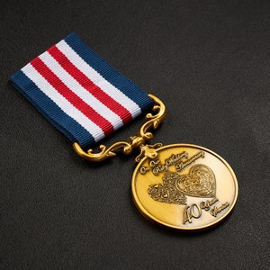 Puede incluir: Una medalla de oro con una cinta roja, blanca y azul. La medalla presenta un diseño de corazón con el texto "On Our Ruby Wedding Anniversary 40 Years Service".